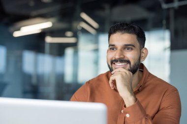 Genç Hintli iş adamı gülümsüyor ve kafasını çeviriyor. Modern ofis ortamında dizüstü bilgisayarla masasında rahatça oturuyor.