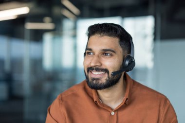 Hintli adam kulaklık takarken gülümsüyor, modern ofis ortamında çalışıyor, mükemmel müşteri hizmetleri ve profesyonel teknik destek sağlıyor.