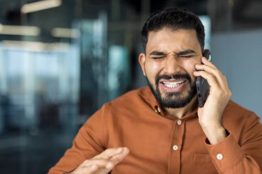 Cep telefonuyla ilgili stresli haberler alan genç adam, modern ofis ortamında iletişim kurarken hayal kırıklığı ve hayal kırıklığı yaşıyor.