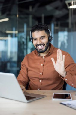 Kulaklık takan genç işadamı, modern ofis ortamında uzaktan kumandalı bir toplantı sırasında dizüstü bilgisayarla çevrimiçi video konferansına katılıyor, gülümsüyor ve el sallıyor.