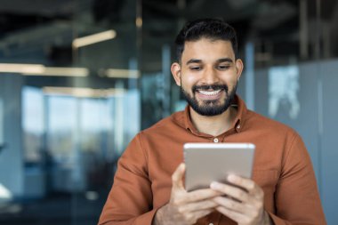 Modern bir ofisteki genç Hintli işadamı dijital tablet kullanırken gülümsüyor, teknoloji odaklı profesyonel iletişim, üretkenlik ve kendine güvenen işyeri işbirliği sergiliyor
