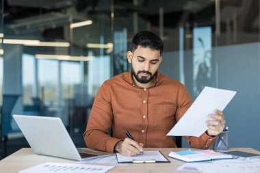 İşadamı, modern bir ofis masasında bilgisayar ve evrak ile sözleşme belgelerini inceleyip imzalamaya, profesyonel sorumluluk, müzakere ve kariyere odaklı kararlılığı aktarmaya odaklandı.
