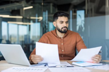 Genç Hindistanlı işadamı modern bir masadaki finansal tabloları ve evrakları dikkatle analiz ediyor, şirket finansmanı ve projeleri için raporlar hazırlıyor ve plan yapıyor.