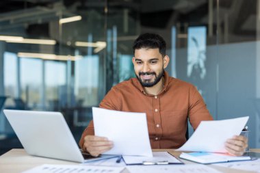 Genç işadamı, ofis masasında mali belgeleri ve raporları inceliyor, evrak işlerini güvenle yürütüyor ve şirket ortamında bir dizüstü bilgisayarla etkili bir şekilde çalışıyor.