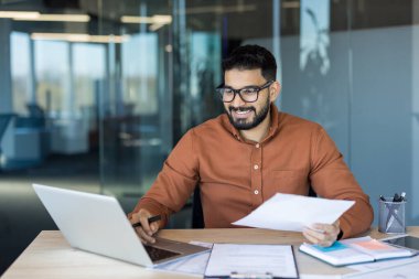 Kendine güvenen Hintli işadamı bilgisayarında çalışıyor ve belgelere gülümseyerek bakıyor. Modern bir ofis ortamında finans ve iş evraklarını yönetiyor.