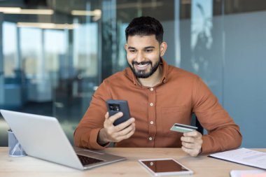 Genç adam elinde akıllı telefon ve kredi kartı tutarken gülümsüyor, çevrimiçi bir ödeme işlerken ya da modern bir iş ortamında ofis masasından alışveriş yaparken.