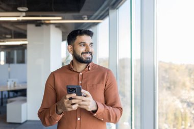 Hintli işadamı gülümsüyor ve elinde akıllı bir telefonla modern bir ofis binasında güvenle dikilirken, uzaktaki pencereden dışarı bakıyor.