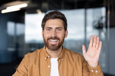 Sanal bir video konferansı sırasında kameraya gülümseyen ve el sallayan bir adam, modern bir ofisten iş arkadaşlarını selamlayan dost canlısı bir iş adamı, uzaktan iletişim için webcam vesikalığı