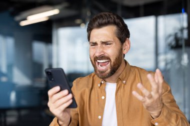 Kahverengi gömlekli öfkeli orta yaşlı bir adam akıllı telefonuna bağırıyor, kötü haberlere ya da stresli internet iletişimine tepki veriyor, öfke, hüsran ve üzgün duygular gösteriyor.