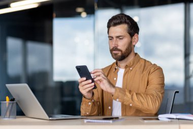 Çalışma odasında oturan sakallı profesyonel bir adam. Yanında bir dizüstü bilgisayar açıkken akıllı telefonuyla uğraşıyor. İş ortamında mobil iletişim ve çoklu görev çizimi