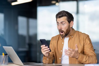 Şaşırmış işadamı ofis masasındaki akıllı telefondan beklenmedik haberler okuyor, bilgisayarları kontrol ederken gözleri ve ağzı açık, şok edici çevrimiçi mesaj veya alarma tepki veriyor.