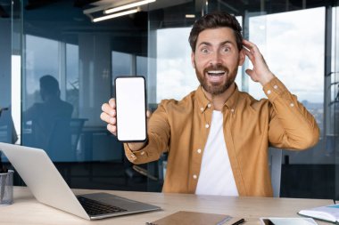 Ofis masasında oturan heyecanlı bir adam, elinde boş beyaz bir ekranla akıllı bir telefon tutuyor, sürpriz ve mutluluğu ifade ediyor, ağzı ve elleri açık bir şekilde kafasına dokunuyor.