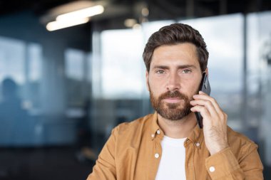 Kulağına akıllı telefon dayamış sakallı bir adam. Çağdaş bir ofis ortamında iletişim ve iş dünyasını temsil ederken ciddi bir telefon görüşmesi yapıyor.