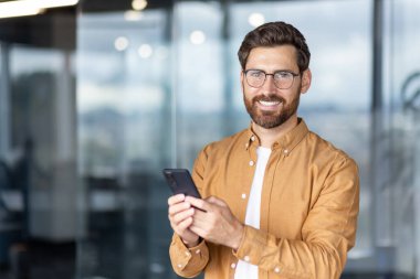 Gözlüklü ve sakallı gülümseyen bir adam cep telefonu kullanıyor, mesajlaşıyor veya internette gezinirken modern ofis ortamında kendine güvenen, bağlantıyı ve dijital iletişimi sembolize eden