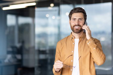 Gülümseyen işadamı başarılı bir telefon görüşmesi yapıyor, modern ofis ortamında göz teması kurarken özgüven ve profesyonelliği ifade ediyor.