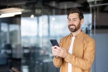 Gülümseyen adam modern bir ofis alanında duruyor, akıllı telefonuyla aktif olarak meşgul oluyor, dijital bağlantısını ve mobil teknolojisini iş ve günlük kullanım için gösteriyor