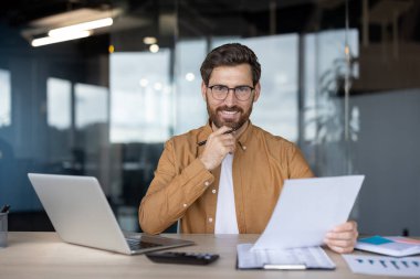 Gözlüklü, gülümseyen iş adamı, modern bir ofiste dizüstü bilgisayarla çalışırken kameraya bakıyor, elinde kalem ve belgeler tutuyordu.