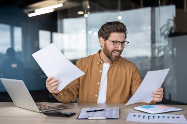 Gözlüklü Kafkas işadamı elinde mali belgeler tutarken şaşkın ve bunalmış görünüyor ve modern ofisindeki bütçe ve vergilerini idare etmeye çalışıyor.