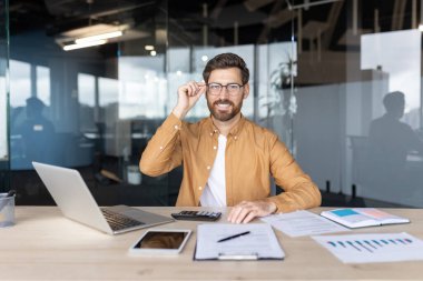 Modern bir ofisteki genç işadamı gözlüğünü ayarlıyor. Masasında dizüstü bilgisayar, tablet, hesap makinesi ve belgelerle çalışırken kameraya gülümsüyor.