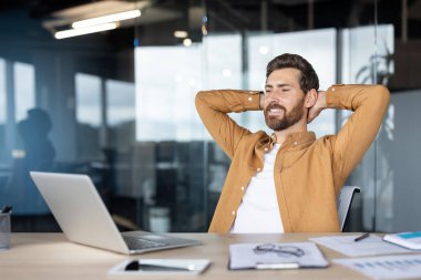 Mutlu genç girişimci işine ara veriyor, elleri başının arkasında sandalyede oturuyor, modern ofis ortamında rahatlığın ve başarının tadını çıkarıyor.