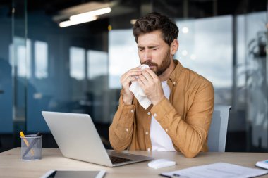Ofis masasındaki bir mendile burnunu sümküren bir adam dizüstü bilgisayar başında çalışırken, hasta görünüyor ve iş günlerinde soğuk ya da mevsimsel alerjilerden yorgun düşüyor.