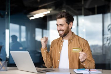 Sakallı adam gülümseyip yumruğunu sıkıyor, elinde altın bir kredi kartı tutarken e-ticaret için bir dizüstü bilgisayar kullanıyor ve başarılı bir online alışveriş yapıyor.