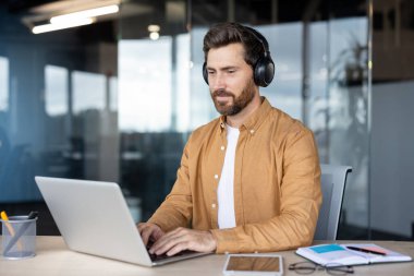 Kablosuz kulaklık takan, bir masada oturan ve dizüstü bilgisayarda yazan, modern ofis ortamında çalışmalarına odaklanan konsantre bir adam.