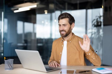 İş adamı gülümseyip el sallarken bilgisayarında sanal bir video görüşmesi yapıyor, modern ofis ortamından uzaktan veya çevrimiçi iletişim kuruyor.