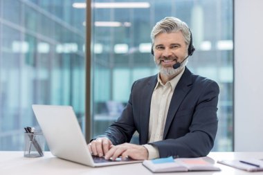 Olgun bir adam kulaklık takıyor ve kameraya gülümsüyor. Modern ofis ortamında dizüstü bilgisayarda yazarken profesyonel müşteri desteği sağlıyor.