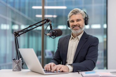 Masada otururken kameraya gülümseyen gri sakallı ve kulaklıklı olgun bir adam. Podcast yayınlamak için dizüstü bilgisayar ve profesyonel mikrofon kullanıyor.