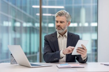 Bir ofis ortamında çalışmak için dijital tablet kullanan bir işadamı, bir dizüstü bilgisayarla bir masada oturuyor ve odaklanmış görünüyor, modern iş iletişimini ve teknoloji kullanımını sembolize ediyor