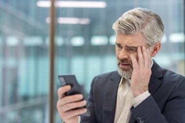 Olgun iş adamı stres ve endişe yaşıyor, akıllı telefondan kötü haberler okuyor, modern ofis ortamındaki zor bir durumdan endişe duyuyor ve şok geçiriyor.