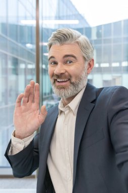 Gri sakallı ve profesyonel giyimli kıdemli işadamı kameraya dostça bir el sallarken neşeli bir şekilde gülümsüyor ve modern ofis ortamında duruyor.