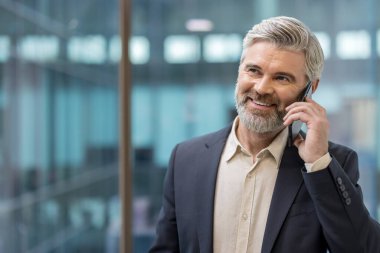Yetişkin kıdemli işadamı cep telefonuyla iletişim kuruyor, şirket ofis ortamında profesyonel bir sohbete girerken güvenle gülümsüyor, başarı ve teknolojiyi temsil ediyor.
