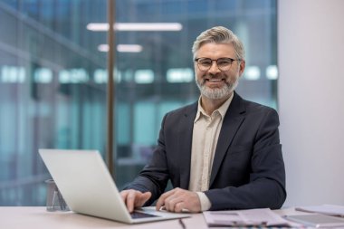 Gri sakallı, gözlüklü, kameraya bakıp gülümseyen, parlak bir şirket ofisinde dizüstü bilgisayarda çalışan olgun bir iş adamı.