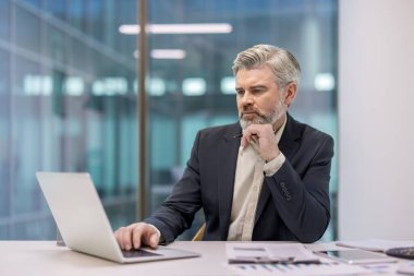 Deneyimli bir işadamı, şirket çağdaş ofisinde masasında oturmuş bilgisayarındaki verileri düşünüyor ve analiz ediyor. Adanmışlığı, stratejiyi, yeniliği ve profesyonel odaklanmayı temsil ediyor.