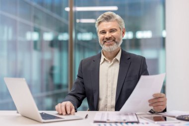 Yetişkin bir işadamı ofis masasında dizüstü bilgisayarla çalışıyor ve belgeleri analiz ediyor. İş başarısı ve kariyeri için profesyonel ve kendinden emin bir imaj çiziyor.