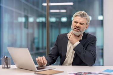 Olgun iş adamı boğazında şiddetli ağrı ve rahatsızlık hissediyor, ofis masasında çalışırken boynunu tutuyor, iş sırasında hastalık ve sağlık sorunlarını gösteriyor.