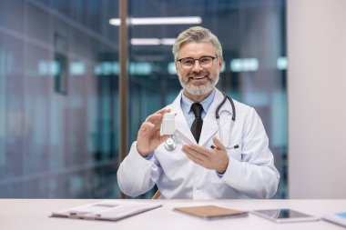 Beyaz önlüklü ve steteskoplu olgun bir erkek doktor elinde beyaz bir ilaç şişesi tutuyor, ilaç sunuyor ve çevrimiçi tele-sağlık danışmanlığı sırasında izleyicilere sağlık tavsiyeleri veriyor.