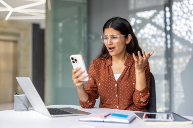 Kadın ofis ortamında akıllı telefonuna bakarken hayal kırıklığı ve stresle tepki veriyor, kötü haberlerle ya da iş günü boyunca bir sorunla uğraşıyor.