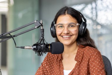 Genç Hintli kadın kulaklıkla güvenle gülümsüyor, dijital medya ve yayın içeriği için podcast veya radyo programı kaydederken stüdyo mikrofonuna konuşuyor.