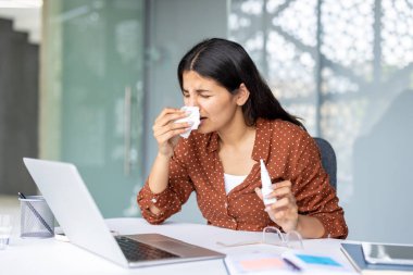 Ofisindeki genç profesyonel kadın soğuk ya da mevsimsel alerjiden muzdarip, burnunu mendile silen ve dizüstü bilgisayarla çalışırken burun spreyi tutan.