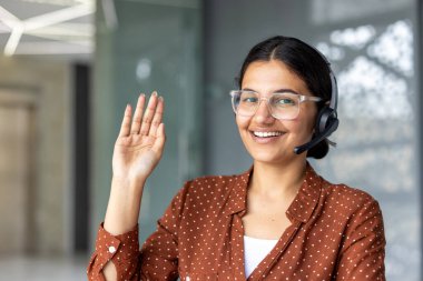 Genç Hintli kadın gözlük ve kulaklık takıyor, neşeli bir şekilde gülümsüyor ve elini sallıyor, modern ofis ortamında online müşteri desteği veya sanal yardım sağlıyor.