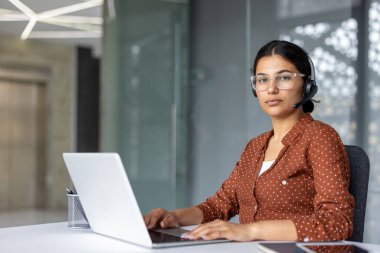 Kulaklık ve gözlük takan kendine güvenen genç bir kadın, beyaz bir ofis masasında oturuyor, gümüş bir dizüstü bilgisayar üzerinde çalışıyor, modern bir çağrı merkezinde müşteri hizmetleri ve destek sağlıyor.