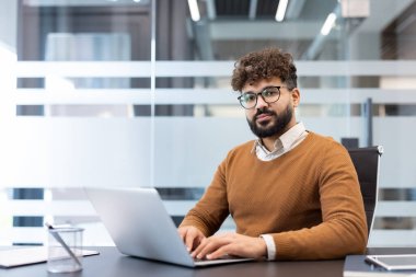 Genç iş adamı birden fazla iş yapıyor, dizüstü bilgisayarda daktilo kullanıyor ve çağdaş bir ofis ortamında izleyiciye güvenle bakıyor, üretkenliği ve profesyonelliği somutlaştırıyor.