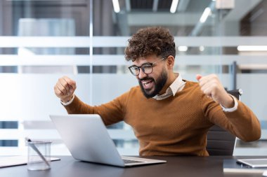 Genç işadamı hedeflerine ulaştıktan sonra zafer kazanıyor, yumruklarını sıkarak dizüstü ekrana bakıyor, ofis ortamında profesyonel zaferi kutluyor ve heyecanlanıyor.