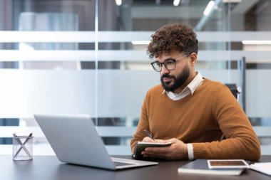 Kıvırcık saçlı ve sakallı bir genç, gözlüklü, bilgisayarından öğrenmeye odaklanmış, modern bir şirket çalışma alanındaki bir ofis masasında not tutuyor.