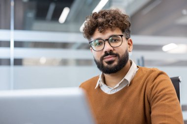 Kıvırcık saçlı ve sakallı genç adam, kameraya özgüvenle bakıyor, modern bir ofiste dizüstü bilgisayarı üzerinde çalışırken, profesyonelliği ve verimliliği temsil ediyor.