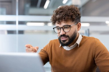 Gözlüklü ve sakallı genç bir adam uzaktan kumandalı bilgisayar ekranına odaklanmış, sanal bir online video görüşmesine katılıyor.