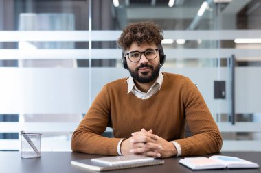 Kulaklık ve gözlük takan genç bir adam, bir ofis masasında oturuyor, bir video görüşmesine katılıyor veya müşteri hizmetleri sunuyor, uzaktan çalışma, destek veya çevrimiçi iletişimi temsil ediyor.